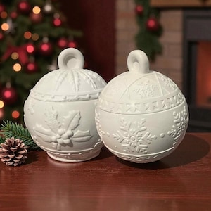 May include: Two white ceramic Christmas bauble-shaped containers with lids. One features a holly design, the other snowflakes. Each has a loop handle. The containers sit on a wooden surface with a Christmas tree and fireplace in the background.