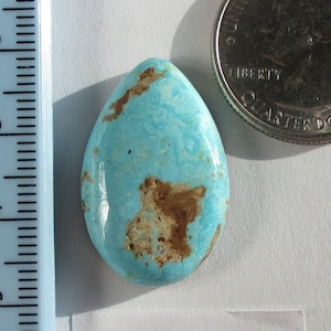 Pode incluir: Uma pedra preciosa turquesa em cabochão com veios marrons e uma cor azul claro. A pedra tem aproximadamente 25,40 quilates e está etiquetada como "Natural Tyrone FL-005-T 25.40ct."