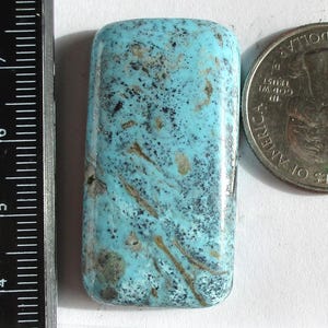 May include: A rectangular, stabilized Kingman turquoise gemstone with a light blue hue and brown matrix patterns. A US quarter coin is next to the gemstone for size comparison. The text "Stabilized Kingman 1HK-61-K 64.3ct." is printed on a white label.