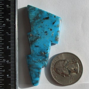May include: A stabilized turquoise stone in the shape of a lightning bolt. The stone is a bright blue color with black veins running through it. The stone is 66.6 carats and is labeled "Kingman Lightning Bolts LN-01-K".
