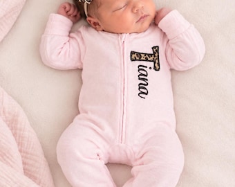 May include: A light pink long-sleeved footed sleeper with a front zipper closure. The name Tiana is embroidered in black script on the chest, with the letter T featuring a leopard print pattern. A matching leopard print bow headband.