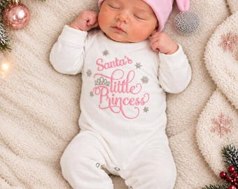 May include: A white long-sleeved baby romper with the pink embroidered text Santa's little Princess and silver snowflake accents, paired with a matching pink beanie featuring the embroidered name Claire and a silver pom-pom.