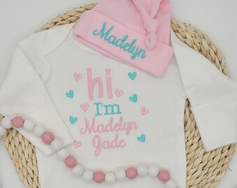 May include: White baby onesie with pink and blue embroidered hearts and the text "hi I'm Madelyn Jade". A pink hat with the name "Madelyn" embroidered on it is on top of the onesie.