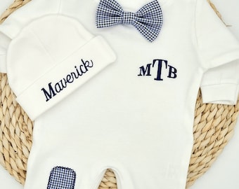 May include: White baby onesie with a blue and white checkered bow tie, a white hat with the name "Maverick" embroidered on it, and the initials "MTB" embroidered on the onesie.