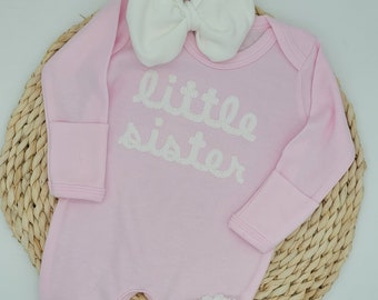 May include: Pink baby onesie with long sleeves and white pearl-like embellishments spelling out "little sister". The onesie has white fabric bow on the neckline and is on a woven straw background.