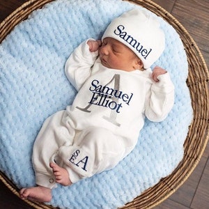 Baby lying in a wicker basket wearing a white coming home outfit romper with a grey last initial and navy first and middle names. Stacked monogram in navy thread on the leg. Navy stitching on a white hat. Customizable names.