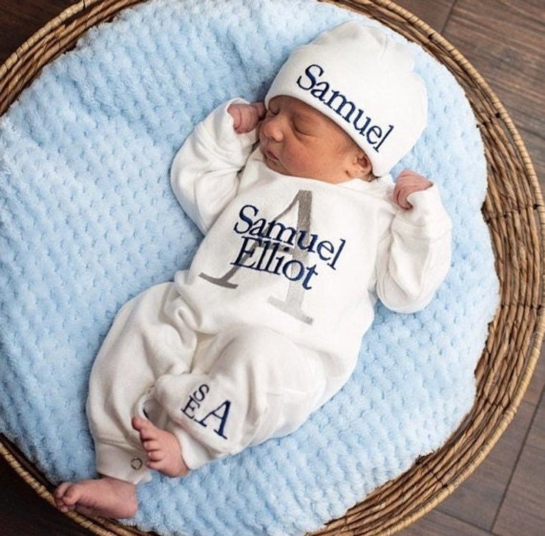 Baby lying in a wicker basket wearing a white coming home outfit romper with a grey last initial and navy first and middle names. Stacked monogram in navy thread on the leg. Navy stitching on a white hat. Customizable names.