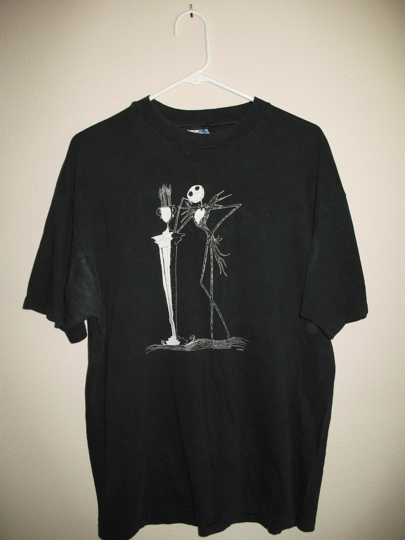 May include: Black t-shirt with a white graphic of Jack Skellington from The Nightmare Before Christmas. The graphic shows Jack holding a cup and standing next to a tall, thin object.