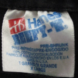 May include: White Hanes tag with blue text that reads "Hanes Beefy-T" and "Pre-Shrunk" in all caps. The tag also includes the text "100% Cotton", "Made in USA", "T Grand X Grande", and "X Large".