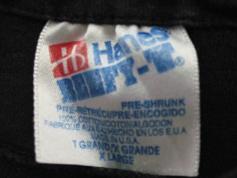 May include: White Hanes tag with blue text that reads "Hanes Beefy-T" and "Pre-Shrunk" in all caps. The tag also includes the text "100% Cotton", "Made in USA", "T Grand X Grande", and "X Large".