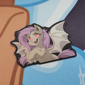 May include: A die-cut sticker depicting a cartoon pony with bat wings, purple hair, and red eyes. The pony has a mischievous grin. The sticker is outlined in black and set against a colourful background.