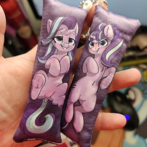 May include: Two purple rectangular keychains featuring cartoon ponies with purple and teal manes. The ponies are light pink with mischievous expressions. The keychains are held together by a silver chain and a white plastic clasp.