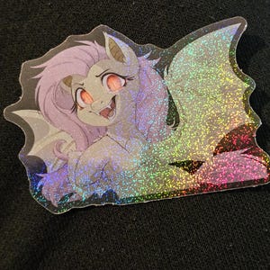 May include: A holographic sticker featuring a cartoon hybrid creature with a horse-like body, bat wings, and a unicorn horn. The creature has pink eyes, purple hair, and a smiling expression. The sticker has a glittery, iridescent finish.
