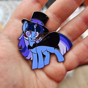 Puede incluir: Un pin de esmalte de pony azul con sombrero de copa, pajarita y cola de arcoíris. El pony tiene pelo morado y lleva un sombrero de copa negro. El pin se sostiene en una mano.