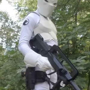 May include: A person wearing a white suit and a white helmet with a black symbol on the left arm, holding a black rifle with a scope.