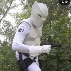 May include: A person wearing a white costume with a white helmet and a black symbol on the chest. The person is holding a black handgun in their right hand. The person is standing in a wooded area.