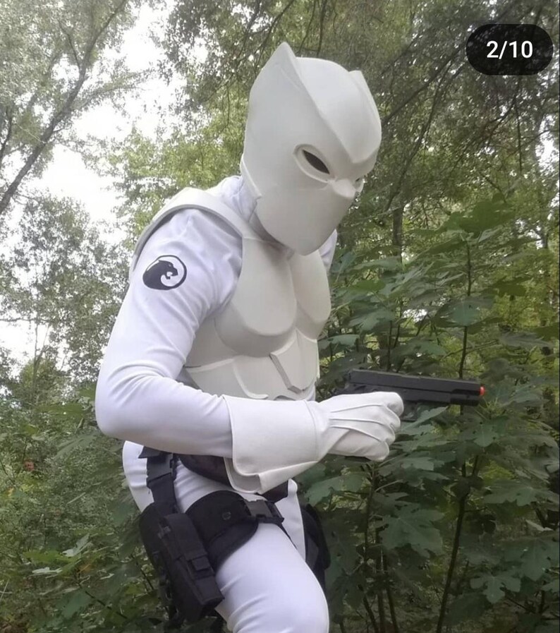 May include: A person wearing a white costume with a white helmet and a black symbol on the chest. The person is holding a black handgun in their right hand. The person is standing in a wooded area.