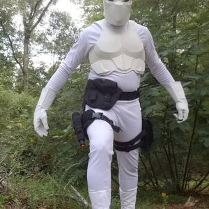 May include: A white costume with a white mask, a black utility belt, and black leg straps. The costume is worn by a person standing in a wooded area.