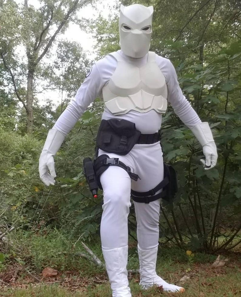 May include: A white costume with a white mask, a black utility belt, and black leg straps. The costume is worn by a person standing in a wooded area.