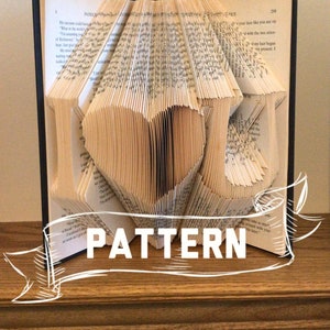 May include: A book folded into the shape of a heart. The book is open and the pages are folded to create a three-dimensional heart shape. The book is on a wooden surface. The text "PATTERN" is written on a white ribbon.