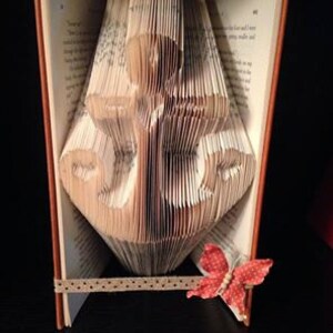 May include: A book with pages folded into the shape of an anchor. The book is standing upright on a table with a brown ribbon and a red butterfly decoration.