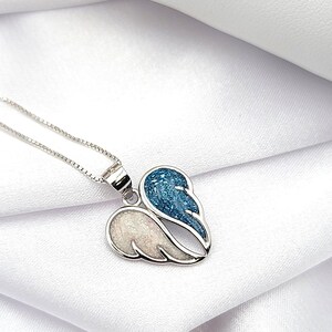 May include: A silver necklace with a heart-shaped pendant featuring two angel wings. One wing is white with glitter, and the other is blue with glitter.