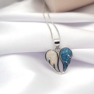 May include: A silver necklace with a heart-shaped pendant featuring two angel wings. One wing is white with a sparkly finish, and the other wing is blue with a sparkly finish.