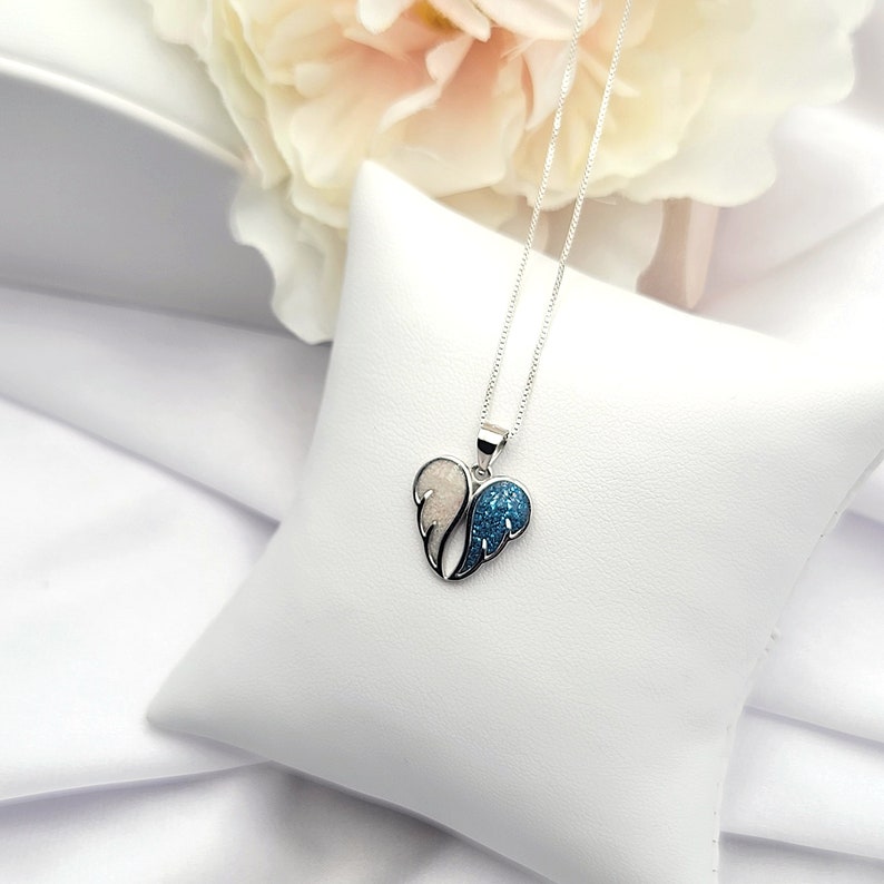May include: A silver necklace with a heart-shaped pendant featuring a blue and white wing design. The wings are covered in sparkly glitter.