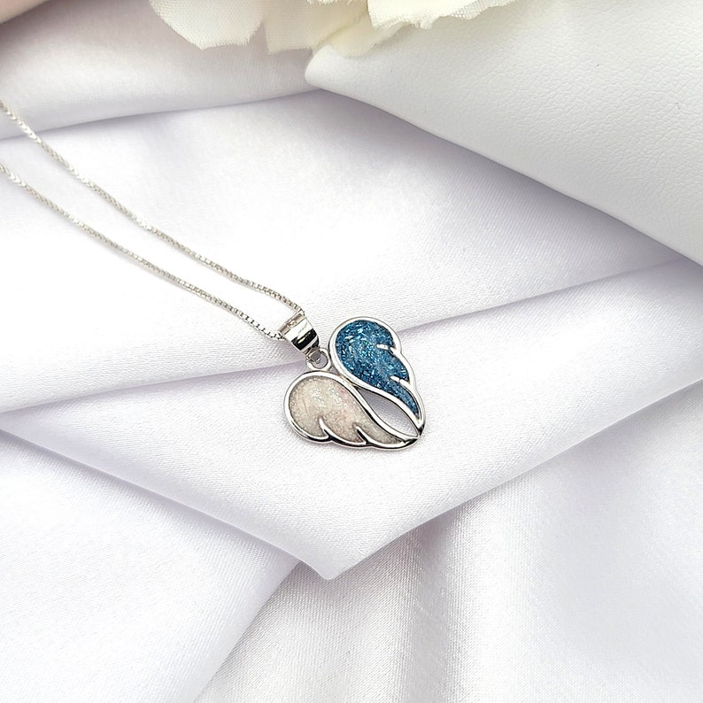 May include: A silver necklace with a heart-shaped pendant featuring two angel wings. One wing is white with glitter, and the other is blue with glitter.