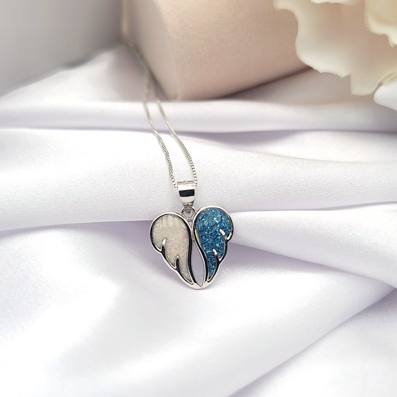 May include: A silver necklace with a heart-shaped pendant featuring two angel wings. One wing is white with a sparkly finish, and the other wing is blue with a sparkly finish.