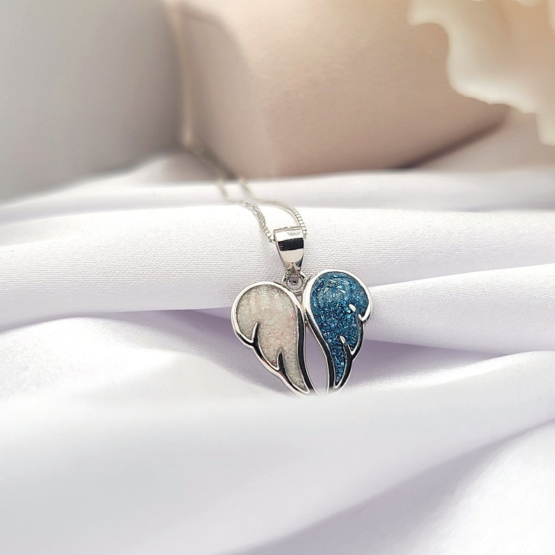 May include: A silver necklace with a pendant featuring two angel wings. One wing is white and the other is blue with glitter.