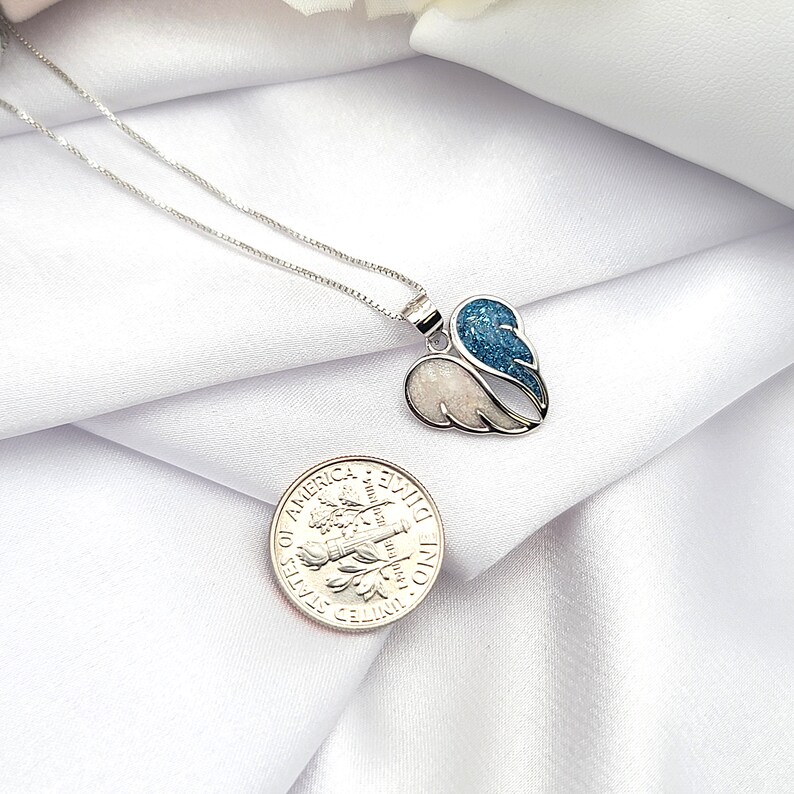 May include: A silver necklace with a heart-shaped pendant featuring two wings. One wing is white and the other is blue with glitter. The pendant is about the size of a US dime.