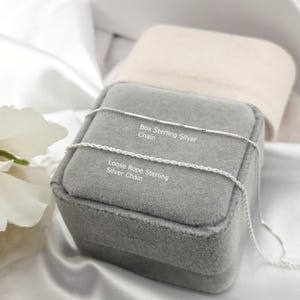 May include: Two silver chains in a gray velvet box. The top chain is a box chain and the bottom chain is a loose rope chain. The text on the box reads "Box Sterling Silver Chain" and "Loose Rope Sterling Silver Chain".