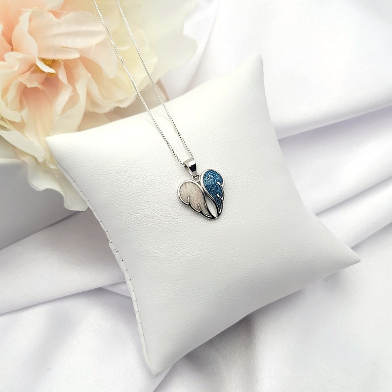 May include: A silver necklace with a heart-shaped pendant featuring two wings. One wing is white and the other is blue with sparkly accents.
