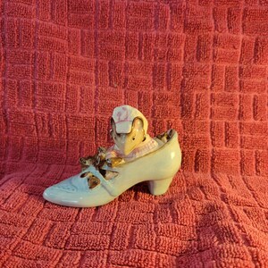 May include: A blue ceramic shoe with a pink and white mouse figurine sitting inside. The mouse is wearing a pink hat and has a pink bow around its neck. There are three smaller mice figurines sitting on the shoe.