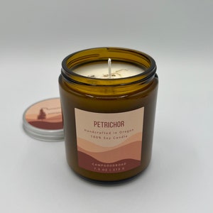 May include: A brown glass candle jar with a white wick and a lid. The label reads "Petrichor, Handcrafted in Oregon, 100% Soy Candle." The candle is 7.5 oz / 213 g. The lid has a landscape design.