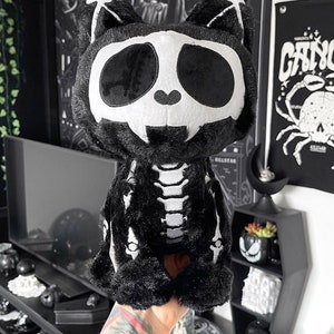May include: Black plush cat skeleton toy with white bones. The cat has a white skull face with black eyes.
