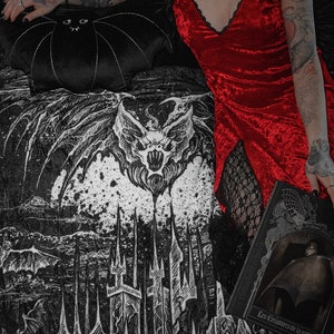 Può includere: Un abito sottoveste rosso in velluto con finiture in pizzo nero è abbinato a una coperta a tema gotico in bianco e nero con un design a pipistrello e castello. Un cuscino nero a forma di pipistrello e un libro intitolato "Les Créatures de la nuit" completano la scena.