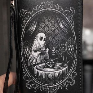 May include: Black wallet with a zipper closure and wrist strap. The front features a black and white illustration of a ghost and a black cat at a table with a skull teapot and cups, inside an ornate oval frame.
