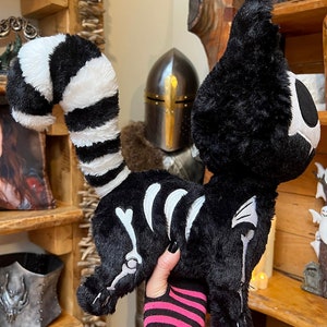 May include: A black and white plush cat with a skeleton design. The cat is being held by a hand wearing a pink and black striped wrist cuff.