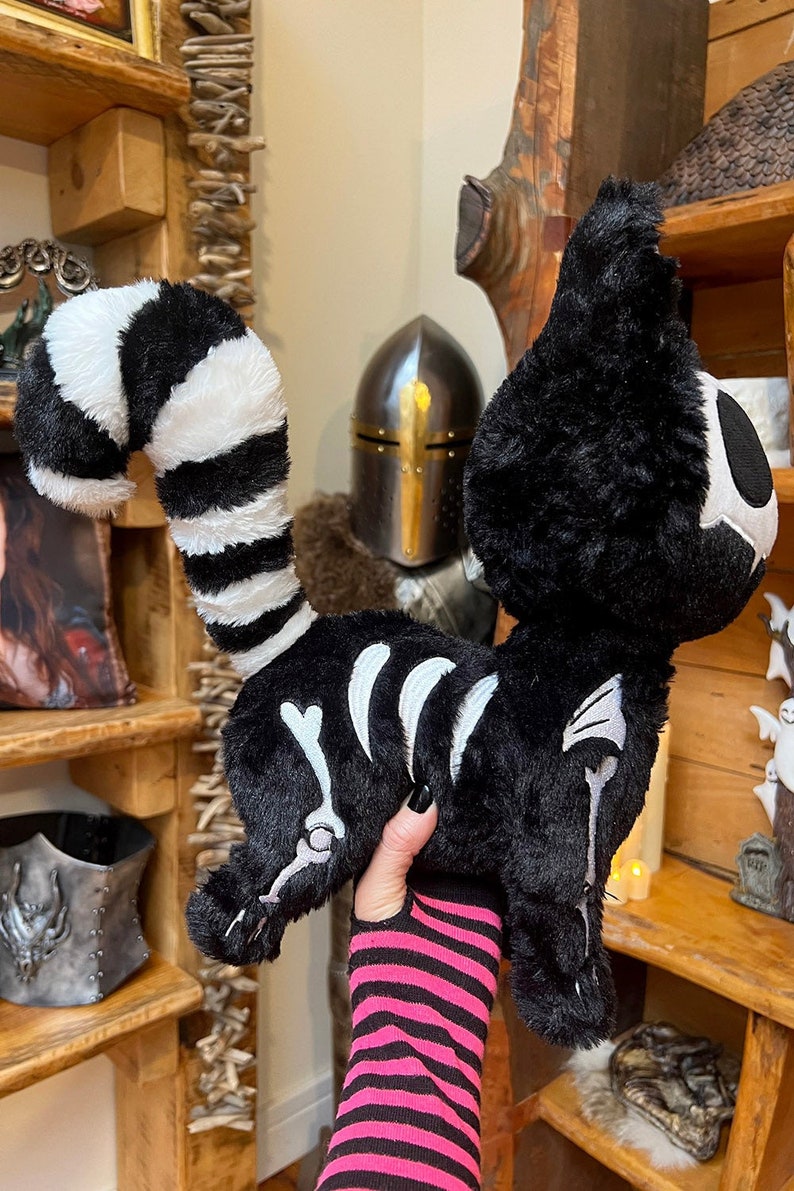 May include: A black and white plush cat with a skeleton design. The cat is being held by a hand wearing a pink and black striped wrist cuff.
