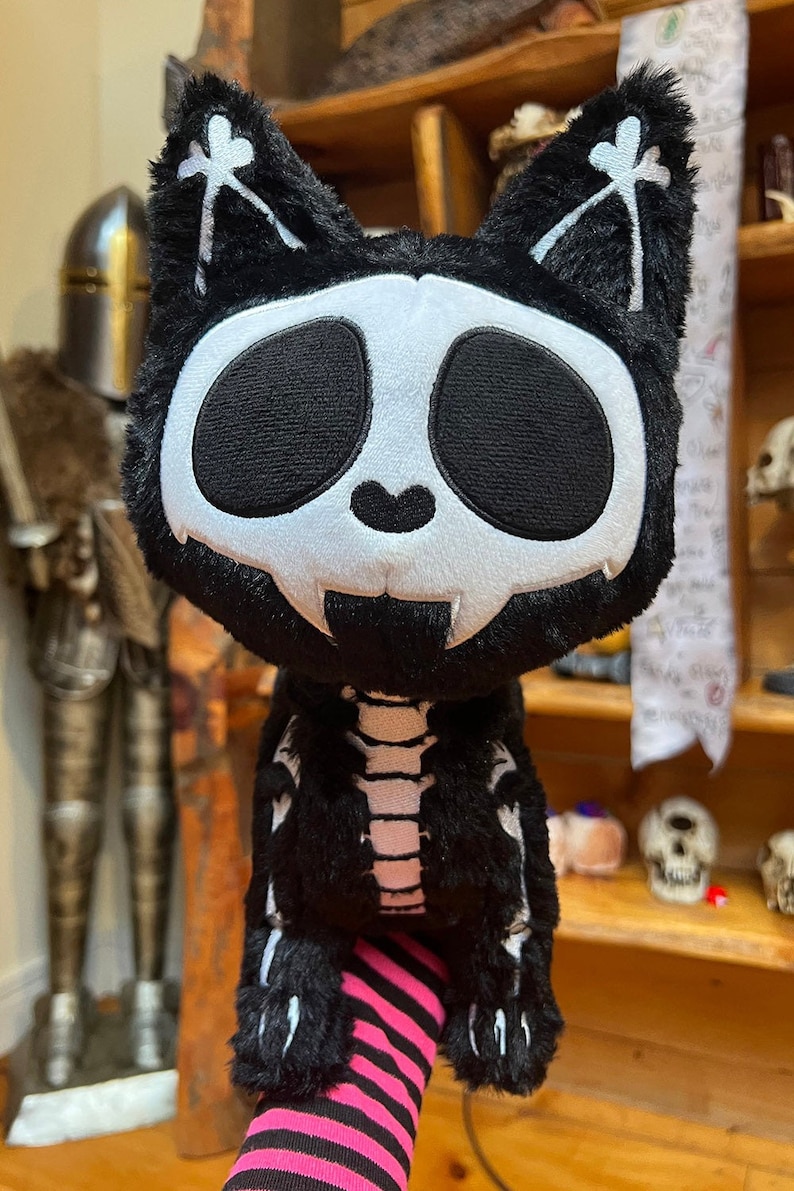 May include: Black plush cat toy with a white skeleton design. The cat has large black eyes and a white skull face. The toy is sitting on a pink and black striped sock.