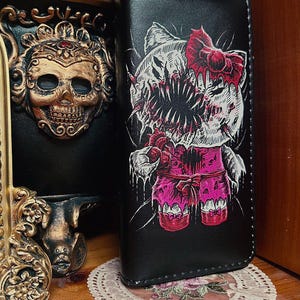 May include: A black rectangular wallet featuring a graphic illustration of a stylized cat character with a large, toothy mouth, a red bow, and a pink outfit. The design includes dripping details and sharp, jagged lines in white, red, and pink ink.