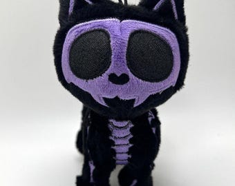 May include: A plush black cat with a purple skull face and skeleton markings. The cat has large black eyes and a small loop for hanging. The plush toy is a unique Halloween or gothic-themed item.