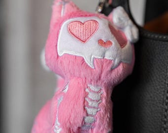 Könnte beinhalten: Ein pinkfarbener Plüsch-Katzen-Schlüsselanhänger mit herzförmigem Gesicht und Skelett-Design. Die Katze hat ein weißes Gesicht mit einem rosa Herzen und einer rosa Nase. Das Skelett-Design ist weiß. Der Schlüsselanhänger ist an einer silbernen Kette befestigt.