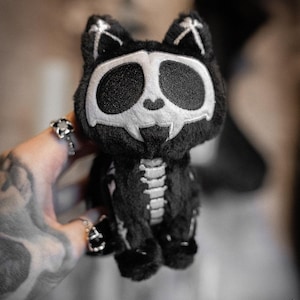 Peut inclure: Peluche noire en forme de chat avec un design de squelette. Le chat a un visage de crâne blanc, une cage thoracique et des détails de colonne vertébrale. Les oreilles ont des os croisés blancs. Un jouet en peluche unique et effrayant.