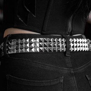May include: A close-up shot of a black belt featuring silver pyramid studs. The belt is worn over black denim jeans. The belt is a fashion accessory.