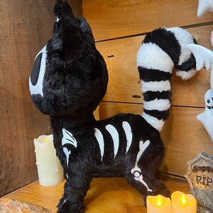 May include: Black plush cat with a white skeleton design and a striped tail. The cat is standing on a wooden shelf with two lit candles.