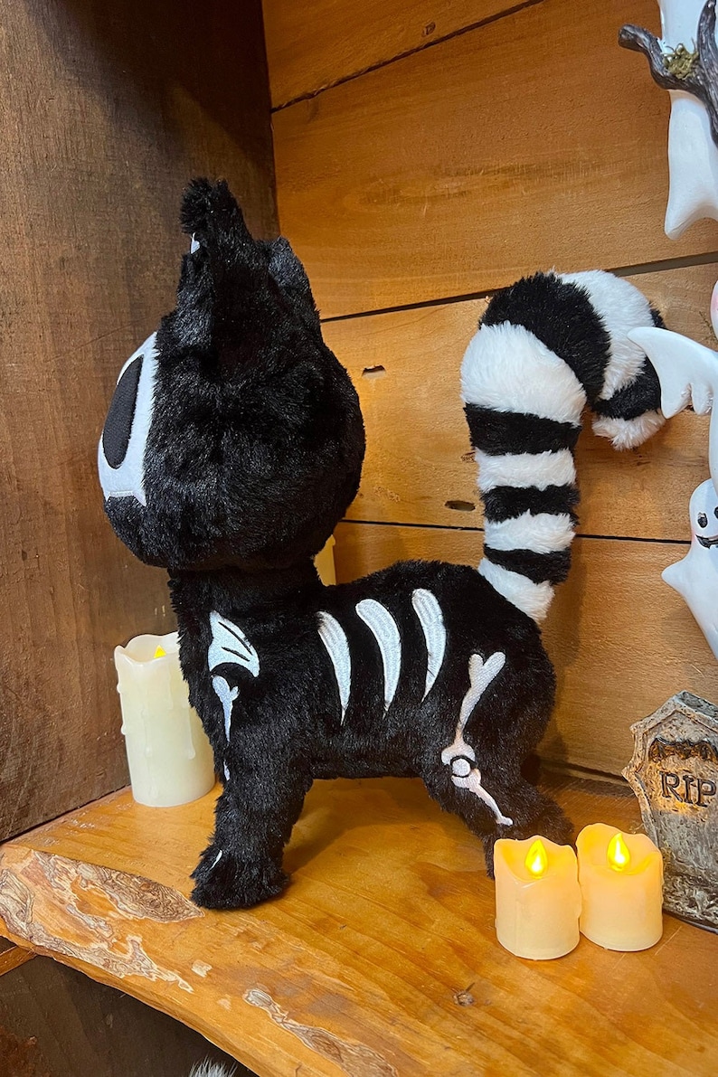 May include: Black plush cat with a white skeleton design and a striped tail. The cat is standing on a wooden shelf with two lit candles.