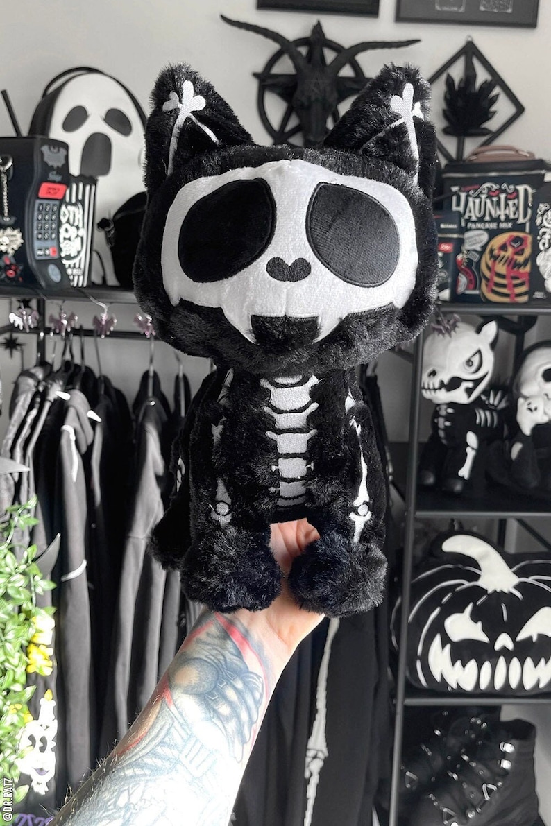 May include: Black plush cat toy with a white skull and skeleton design. The toy is being held by a hand with a tattoo.